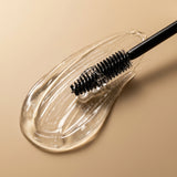 Escovinha aplicadora sobre a textura transparente do Sérum Moon Lashes, em fundo bege, mostrando que a fórmula é incolor e de toque leve.