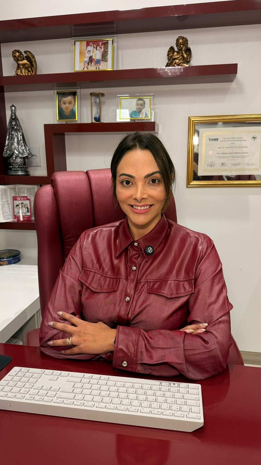 Dra. Fabiana Caetano sentada à mesa do consultório da Imderme, com o título de especialista em dermatologia na parede e retratos de família nas prateleiras