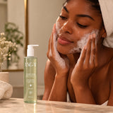 Mulher lavando o rosto com espuma do Sabonete Facial Lunar Glow 300ml da Lagai, frasco com gel verde na bancada do banheiro.